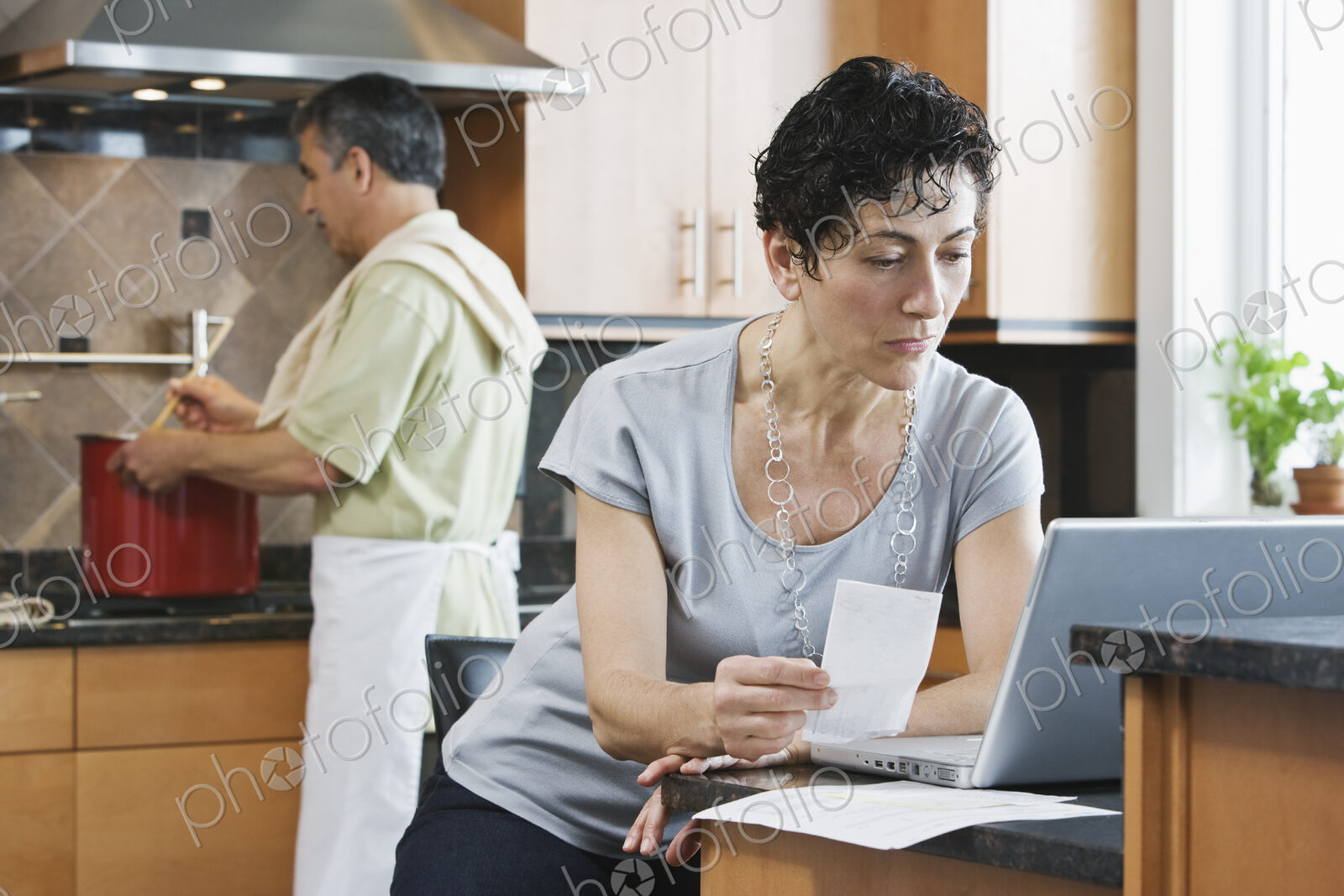 A man cooking supper at the hob, and a woman using a laptop and checking paperwork in…