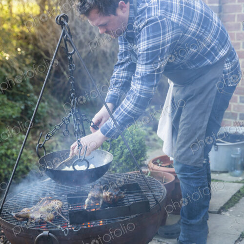 A man cooking vegetables in a pan suspended from a trivet over a glowing fire. A man cooking vegetables in a pan suspended from a trivet over a glowing fire.