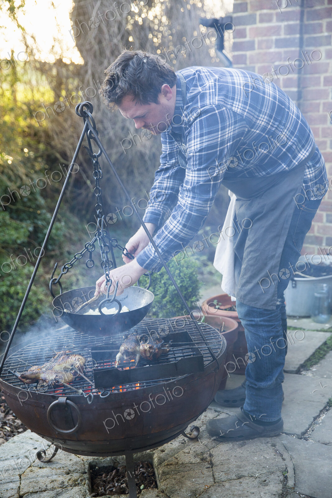 A man cooking vegetables in a pan suspended from a trivet over a glowing fire.