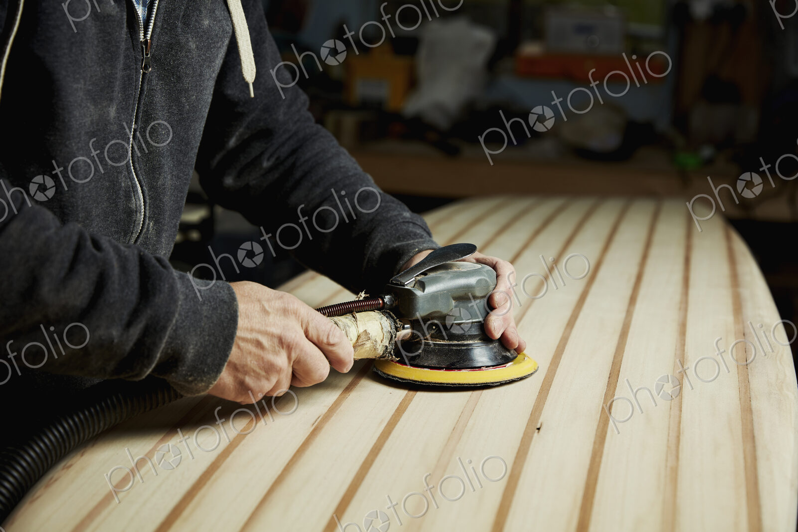 A man finishing the surface of a wooden surfboard with an electrical sander.
