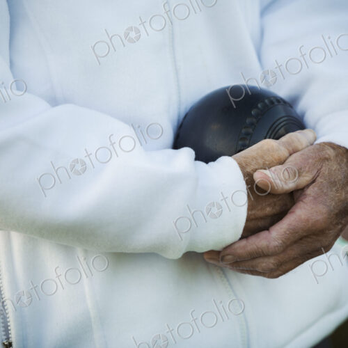 A man holding a black wooden lawn bowls ball in his hands close to his chest. A man holding a black wooden lawn bowls ball in his hands close to his chest.