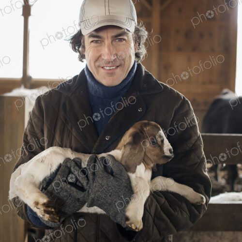 A man holding a small kid goat in his arms in a livestock pen.