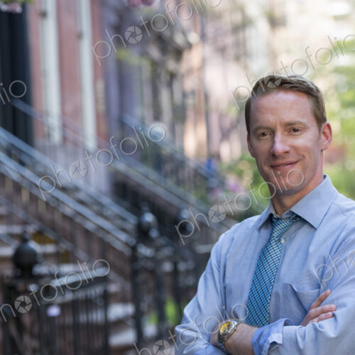 A man in a blue shirt and blue tie with arms folded, standing on the sidewalk outside… A man in a blue shirt and blue tie with arms folded, standing on the sidewalk outside…