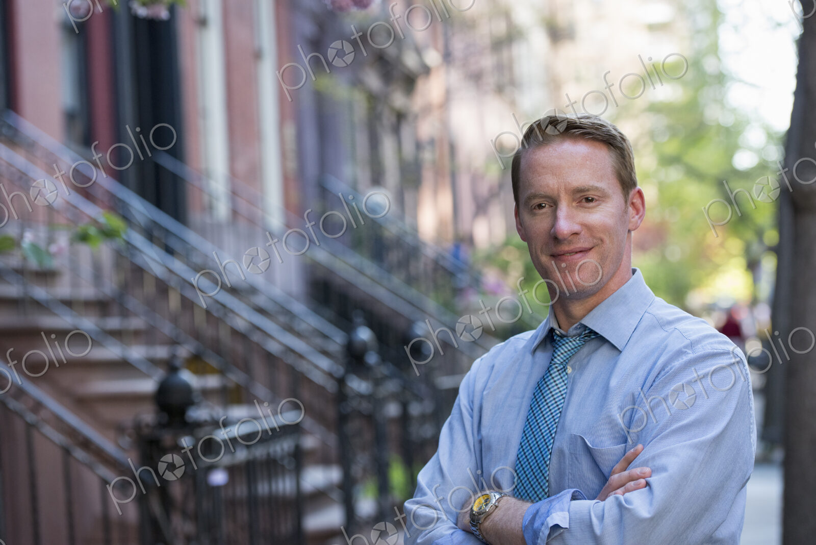 A man in a blue shirt and blue tie with arms folded, standing on the sidewalk outside…