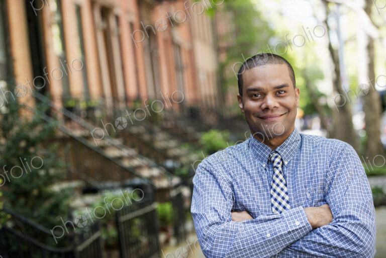 A man in a blue shirt and tie standing on a city street. A terrace of townhouses.