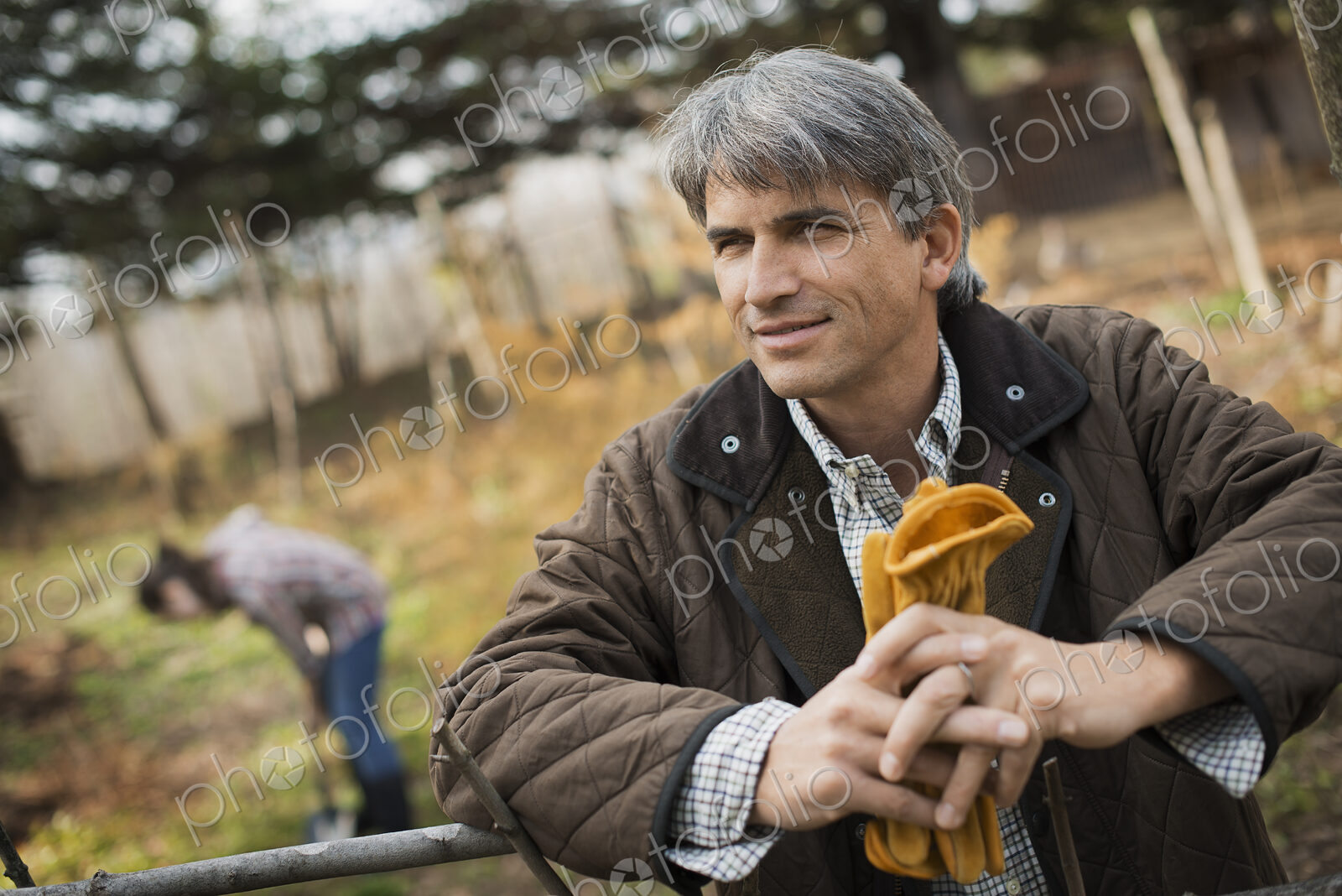 A man in a brown jacket, holding leather work gloves on an organic farm. A person…