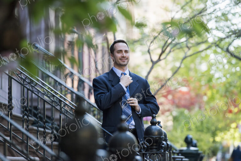 A man in a business suit adjusting his tie. At the bottom of the steps of a townhouse…