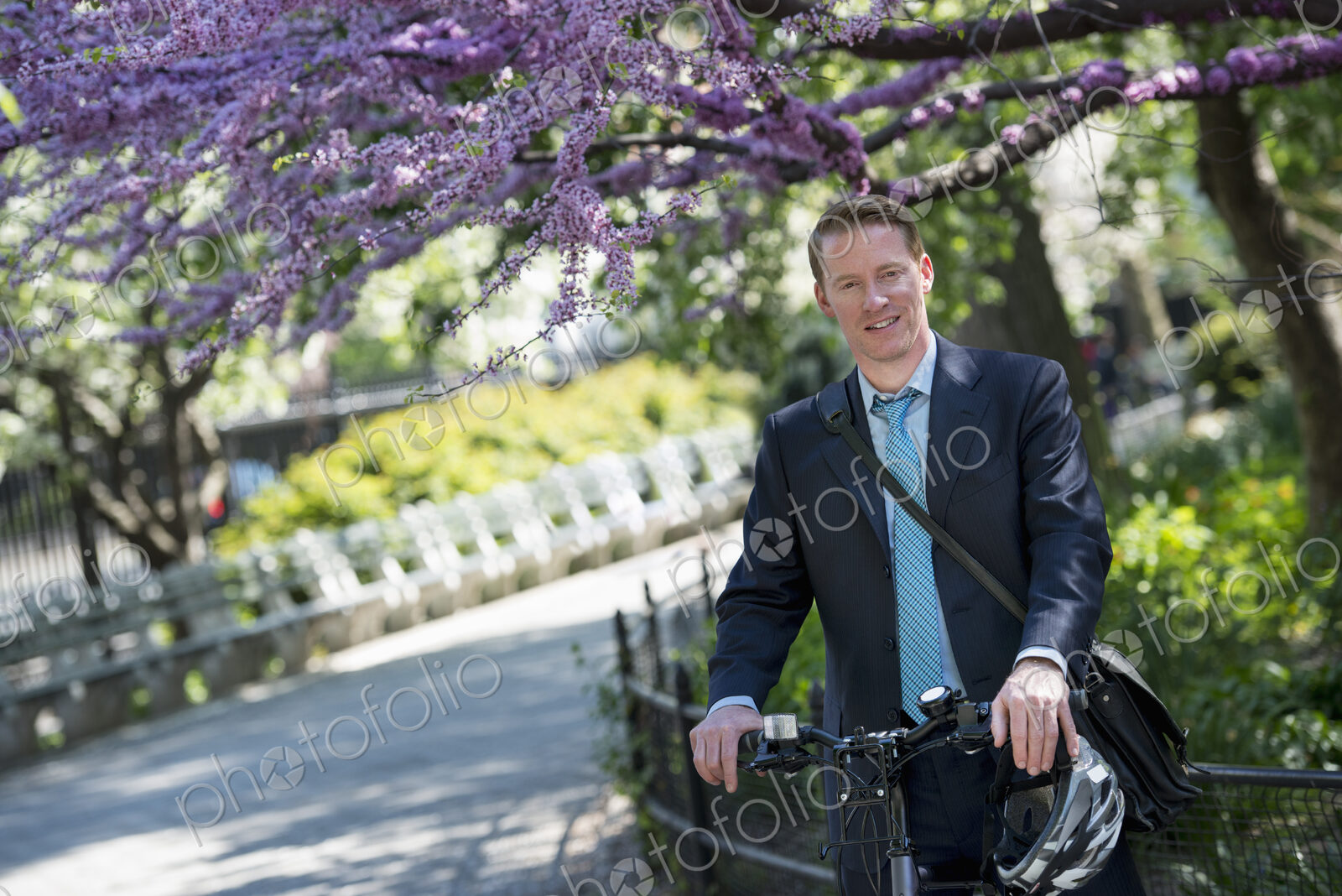 A man in a business suit, outdoors in a park. Sitting on a bicycle, with a cycle…