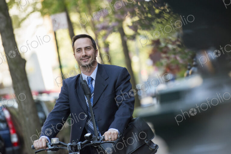 A man in a business suit, outdoors in a park. Sitting on a bicycle, with a cycle…
