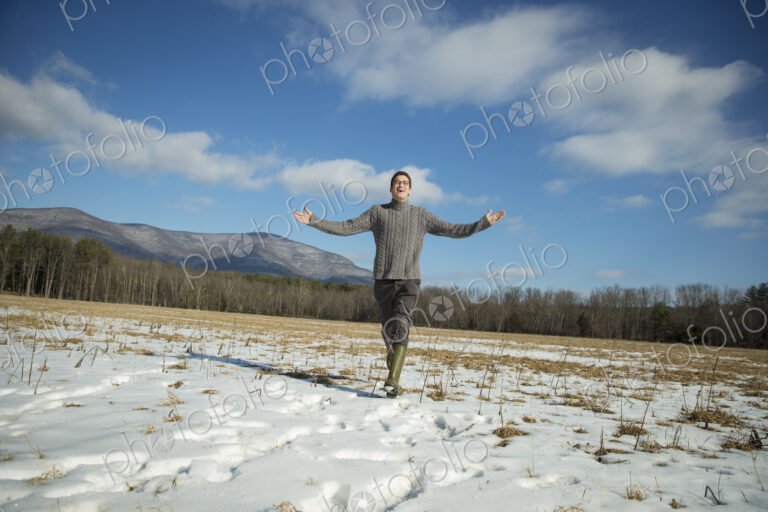 A man in a cable knit jumper and muck boots standing with his arms stretched out, in a…
