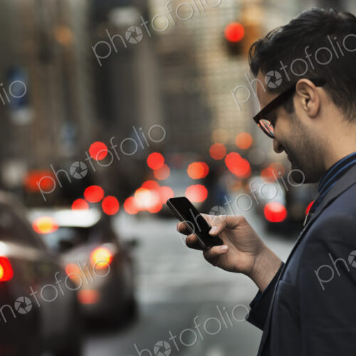 A man in a dark jacket checking his cell phone, standing on a busy street at dusk. A man in a dark jacket checking his cell phone, standing on a busy street at dusk.