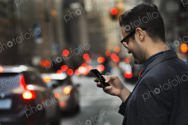 A man in a dark jacket checking his cell phone, standing on a busy street at dusk.