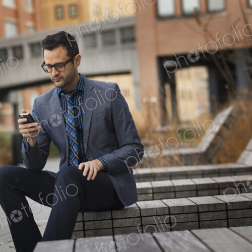 A man in a formal jacket and tie, sitting on a bench outside a city building, checking…