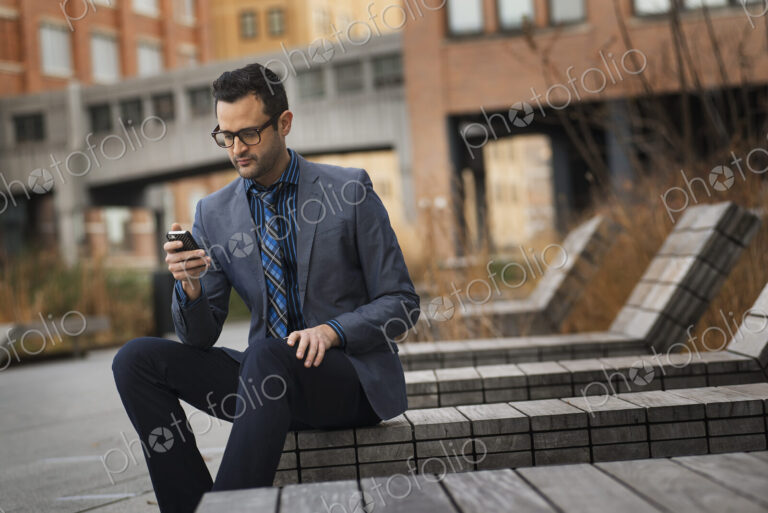 A man in a formal jacket and tie, sitting on a bench outside a city building, checking…