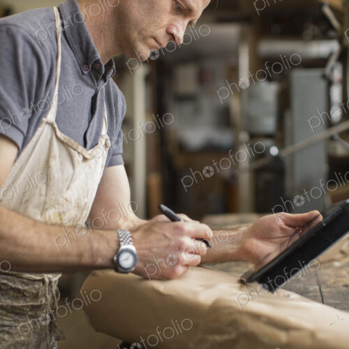 A man in a furniture restoration workshop using a digital tablet, writing on a packet… A man in a furniture restoration workshop using a digital tablet, writing on a packet…
