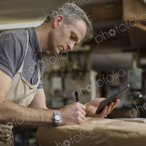 A man in a furniture restoration workshop using a digital tablet, writing on a packet… A man in a furniture restoration workshop using a digital tablet, writing on a packet…