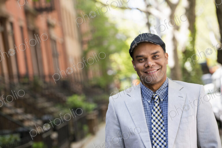 A man in a jacket and tie on a city street outside a terrace of brownstone houses.