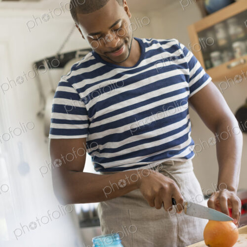 A man in a kitchen using a knife to slice an orange. A man in a kitchen using a knife to slice an orange.