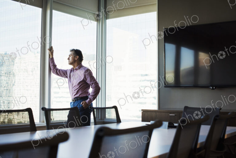 A man in a meeting room looking out of a window at an urban landscape.