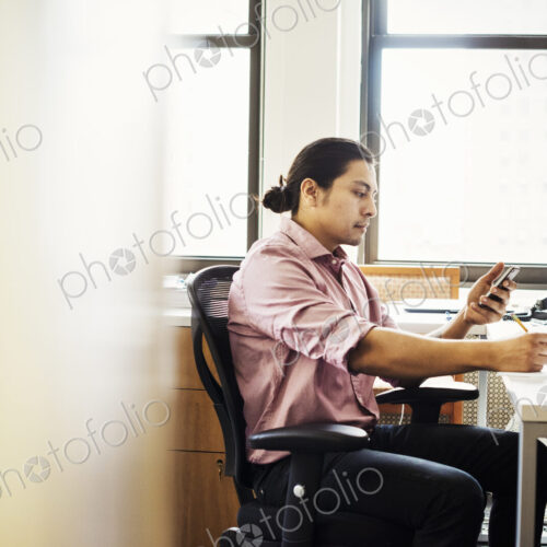 A man in a pink shirt in an office checking his phone, viewed through an open door. A man in a pink shirt in an office checking his phone, viewed through an open door.