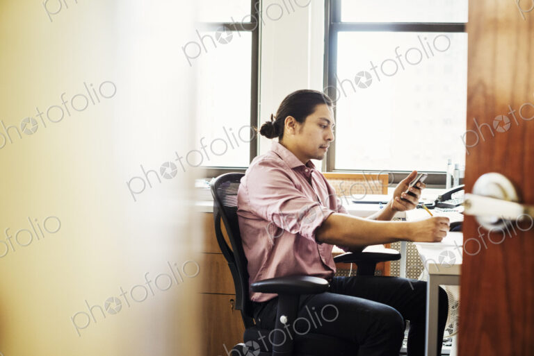 A man in a pink shirt in an office checking his phone, viewed through an open door.