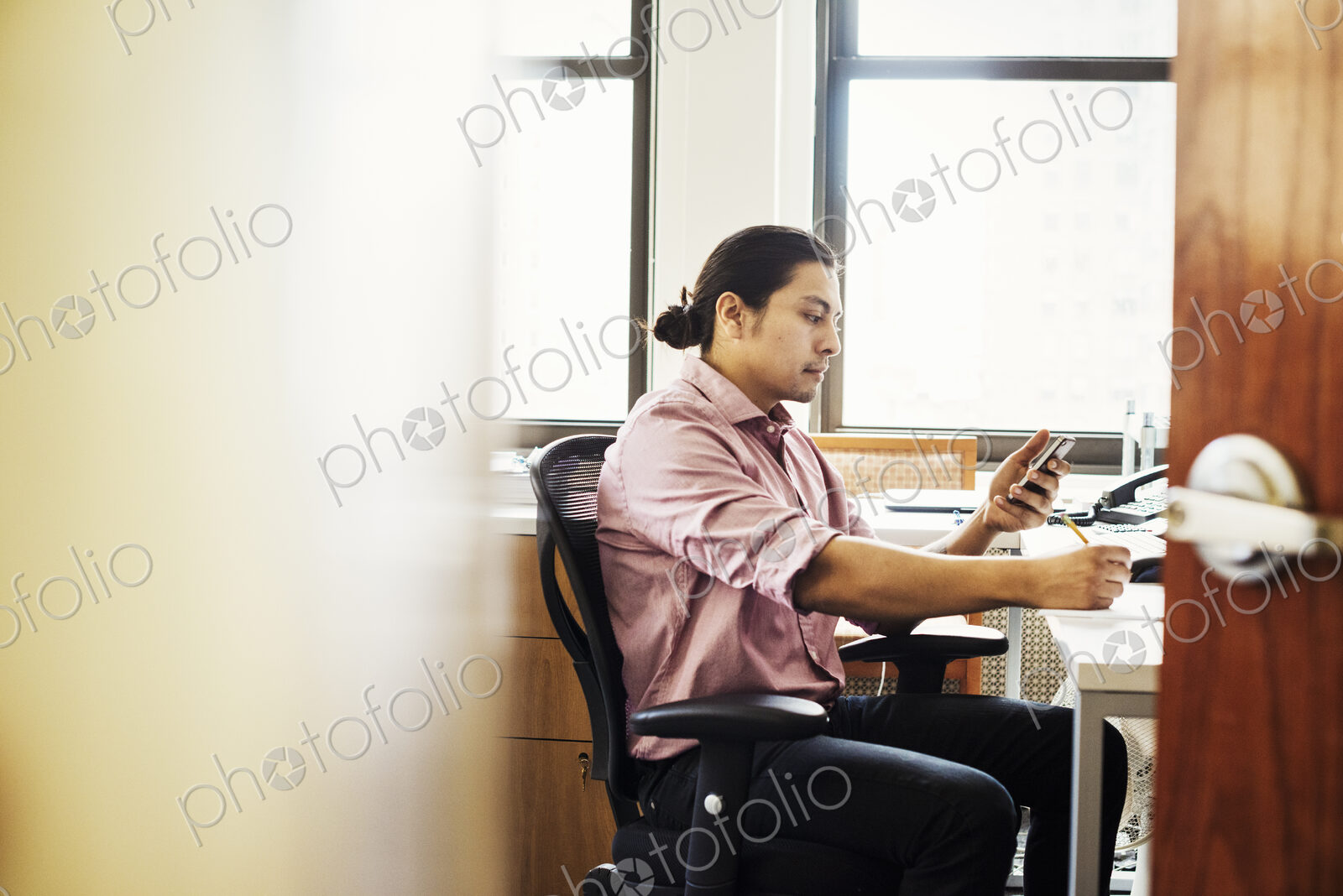 A man in a pink shirt in an office checking his phone, viewed through an open door.