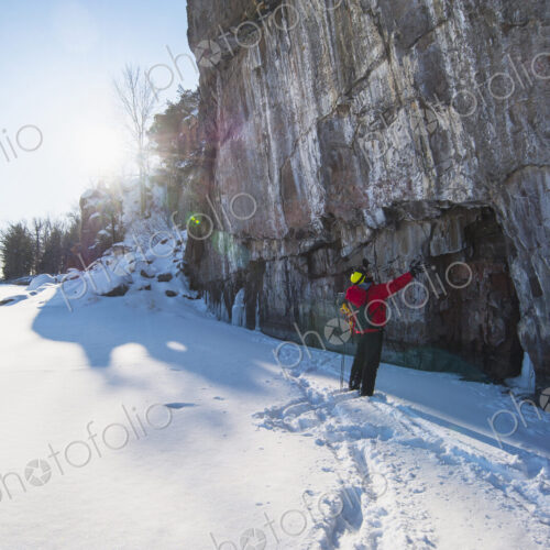 A man in a red jacket standing in front of an ice cliff looking up, ice climbing.