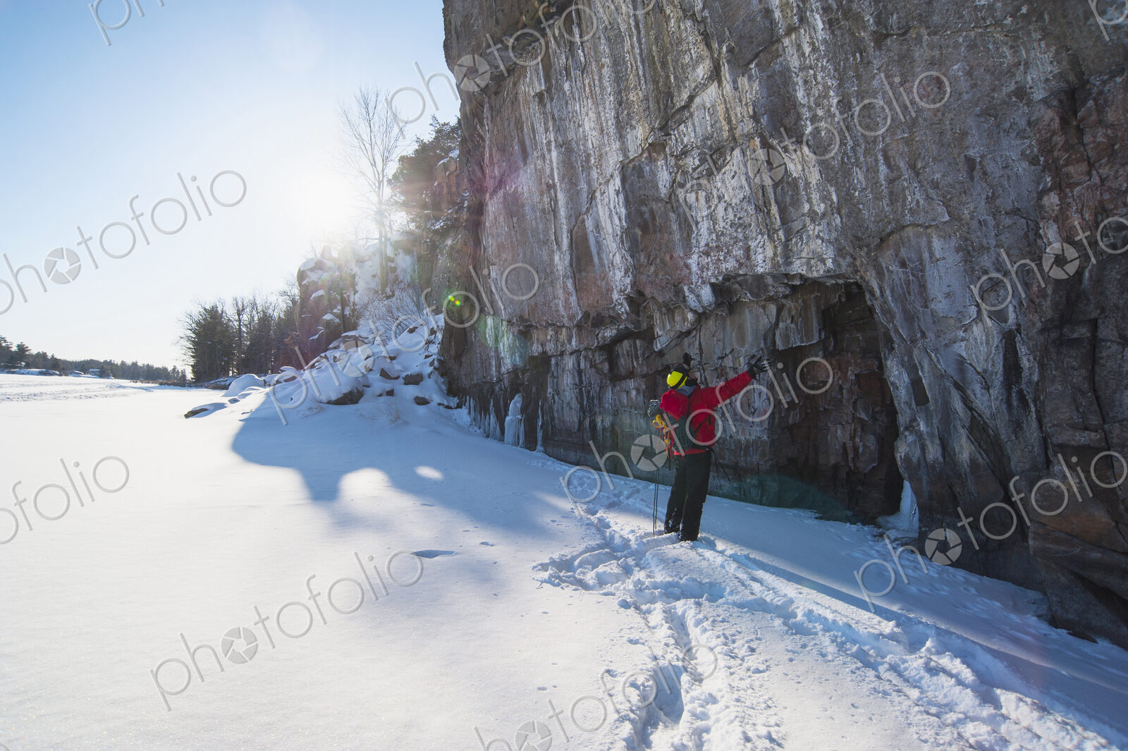 A man in a red jacket standing in front of an ice cliff looking up, ice climbing.