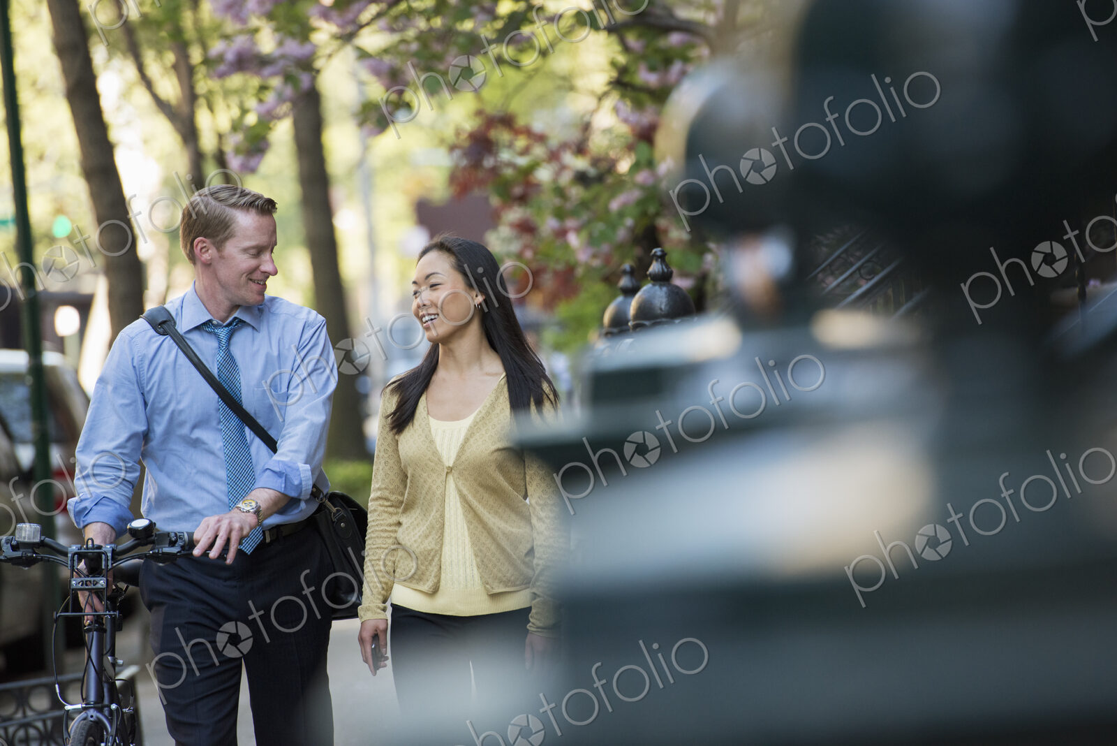 A man in a shirt and tie pushing a bicycle, and talking to a woman. A park in the city.