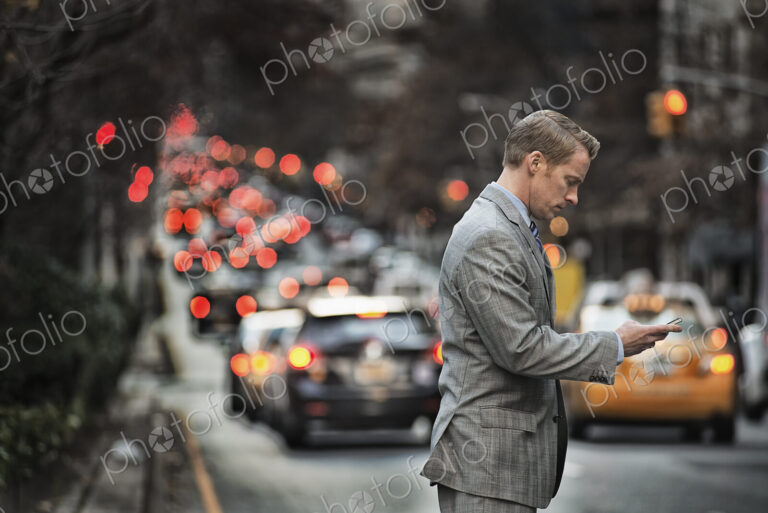 A man in a suit checking his cell phone, standing on a busy street at dusk.