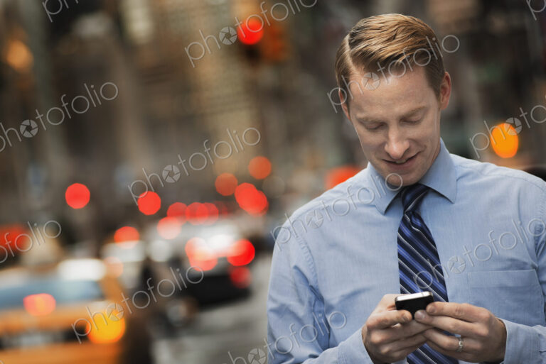 A man in a suit checking his cell phone, standing on a city sidewalk at dusk.