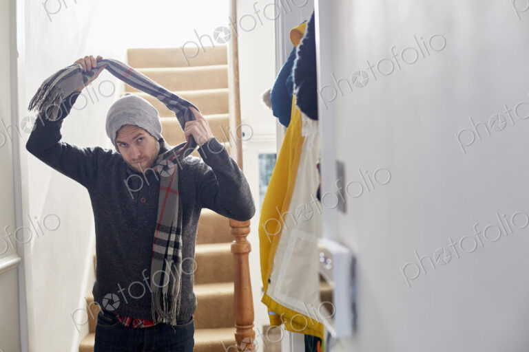A man in a winter coat, hat and scarf arriving home, taking his scarf off.