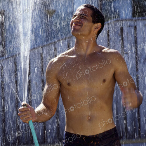 A man in shorts with a bare chest, holding a garden hose and standing in the spray on… A man in shorts with a bare chest, holding a garden hose and standing in the spray on…