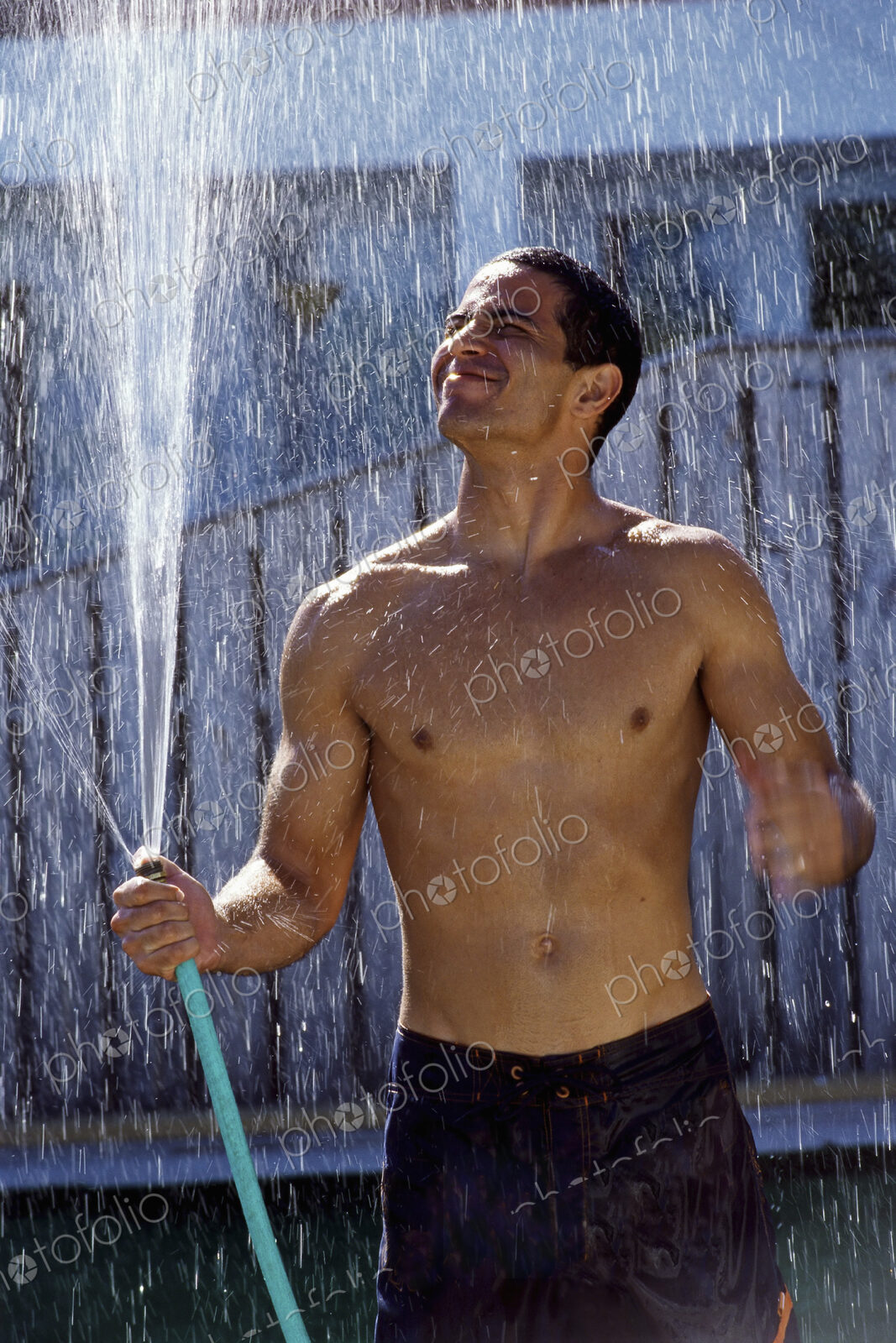 A man in shorts with a bare chest, holding a garden hose and standing in the spray on…