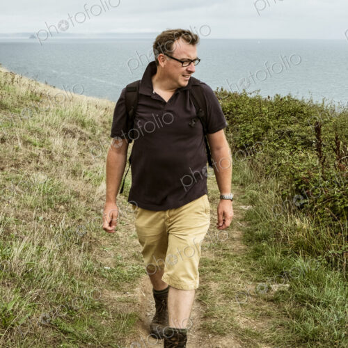 A man in walking boots with a rucksack, walking along a coastal path.