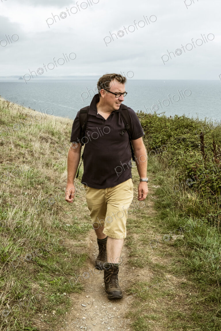 A man in walking boots with a rucksack, walking along a coastal path.