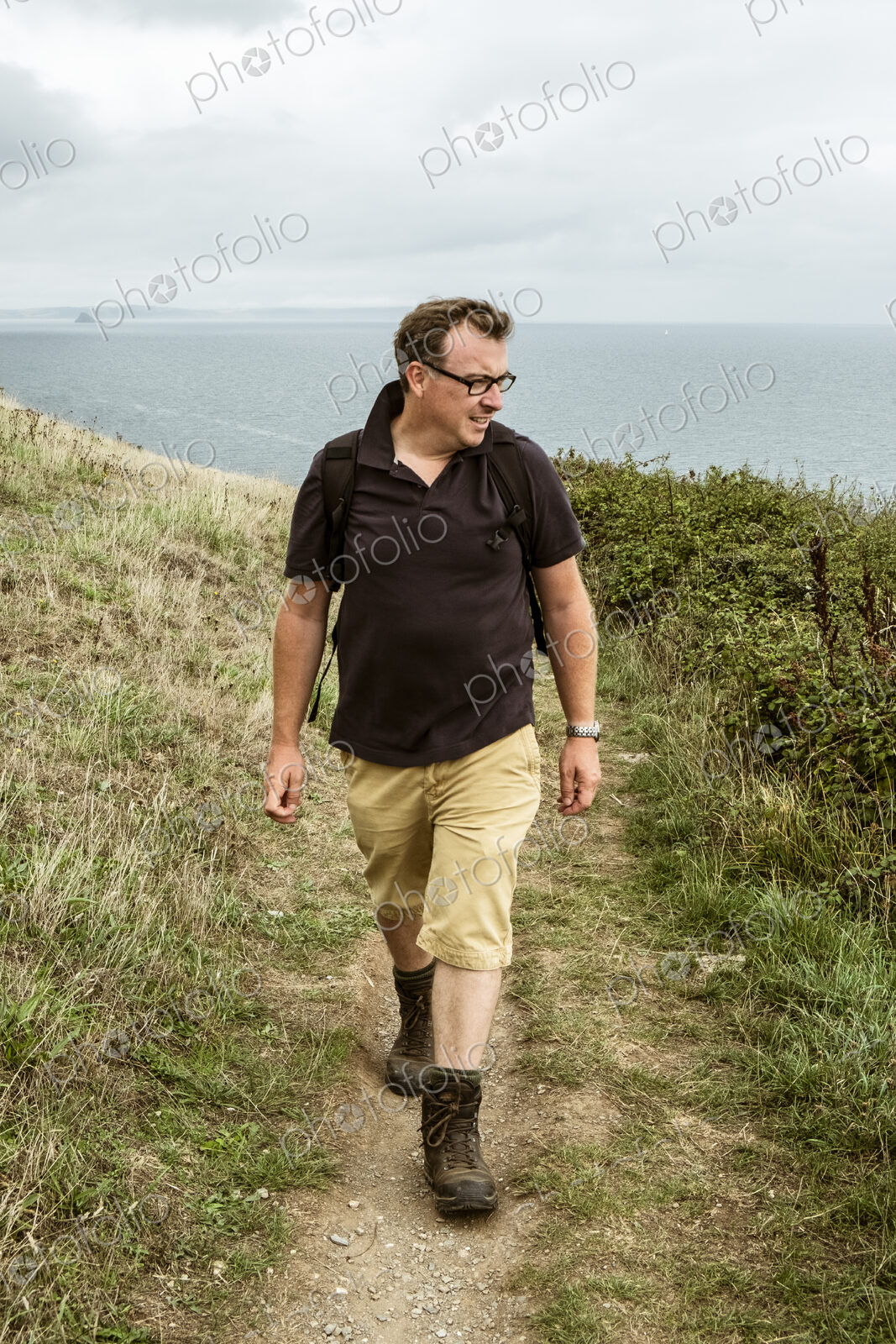 A man in walking boots with a rucksack, walking along a coastal path.