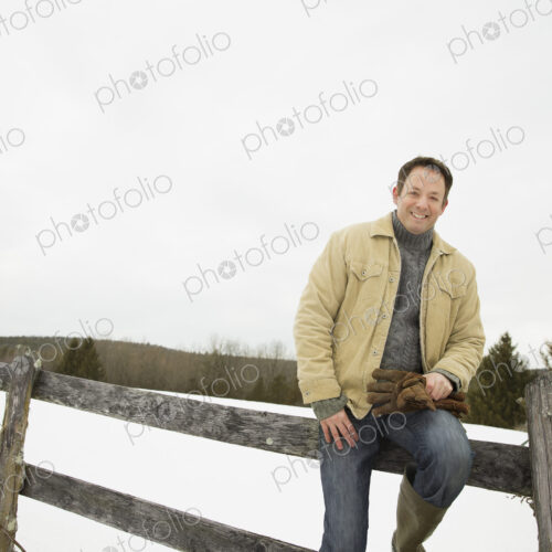 A man in winter jacket with gloves, sitting on a fence in winter snow. A man in winter jacket with gloves, sitting on a fence in winter snow.