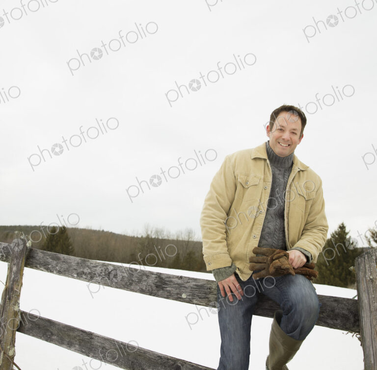 A man in winter jacket with gloves, sitting on a fence in winter snow.
