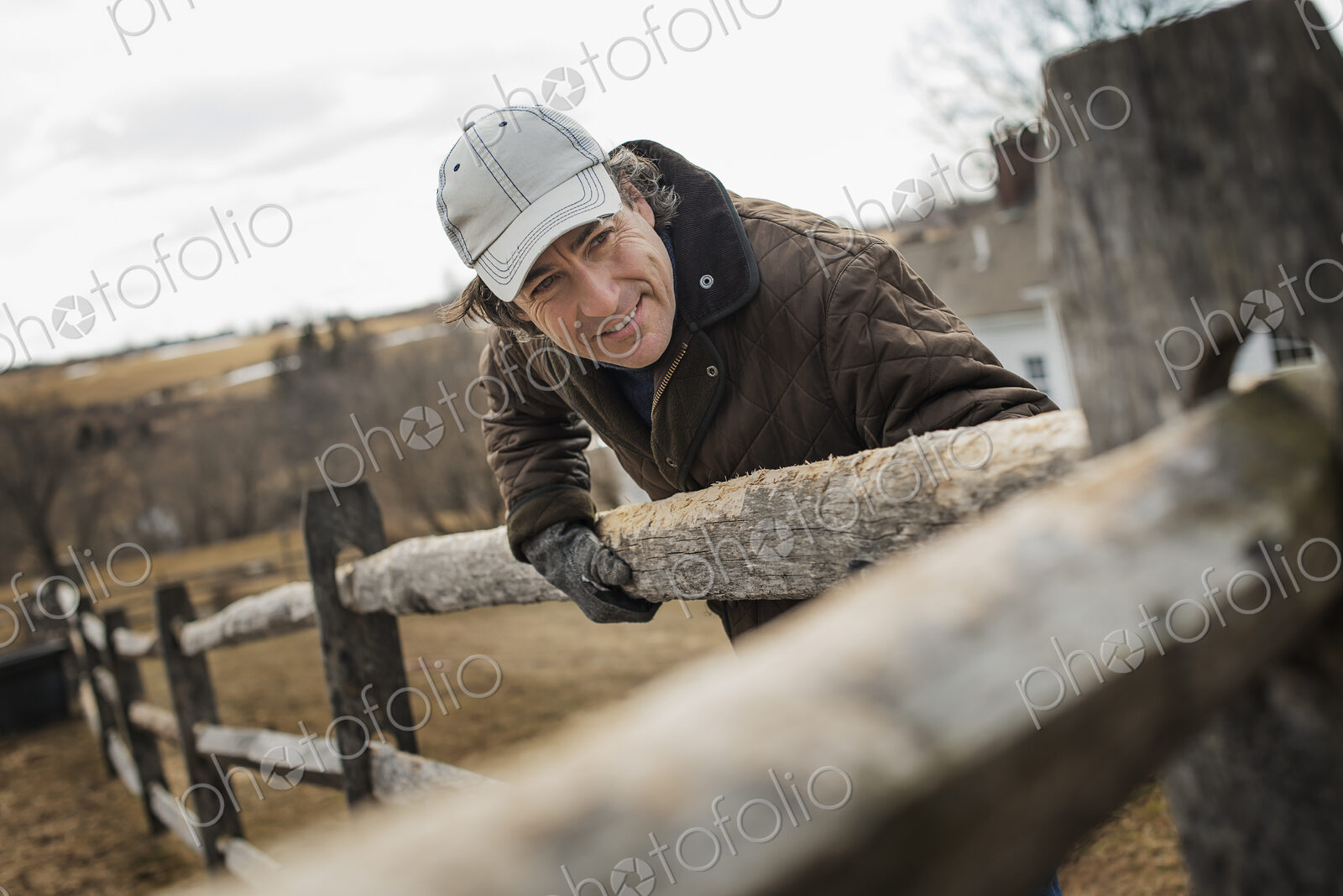 A man leaning against a post and rail fence on a farm in winter.