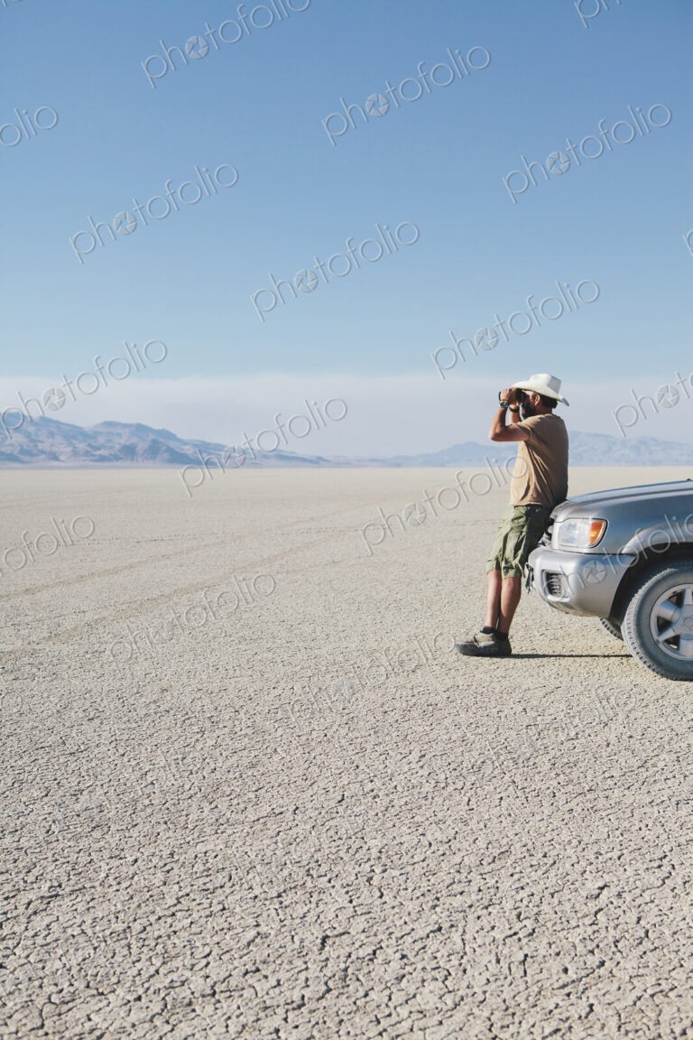 A man leaning against a truck, looking through binoculars.