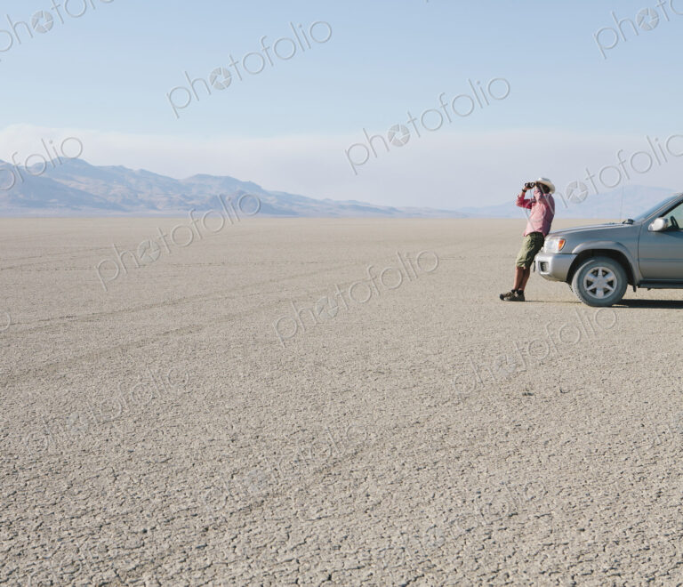 A man leaning against a truck, looking through binoculars.
