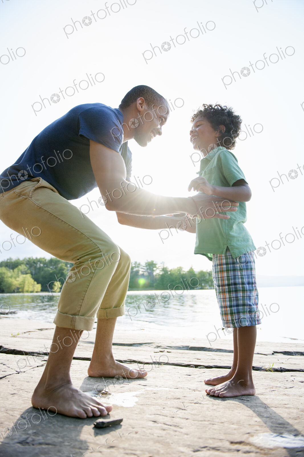 A man lifting a small boy up, playing in the sun by a lake.