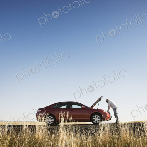 A man looking in the engine compartment of his parked car parked along a highway