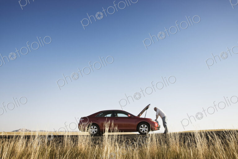 A man looking in the engine compartment of his parked car parked along a highway