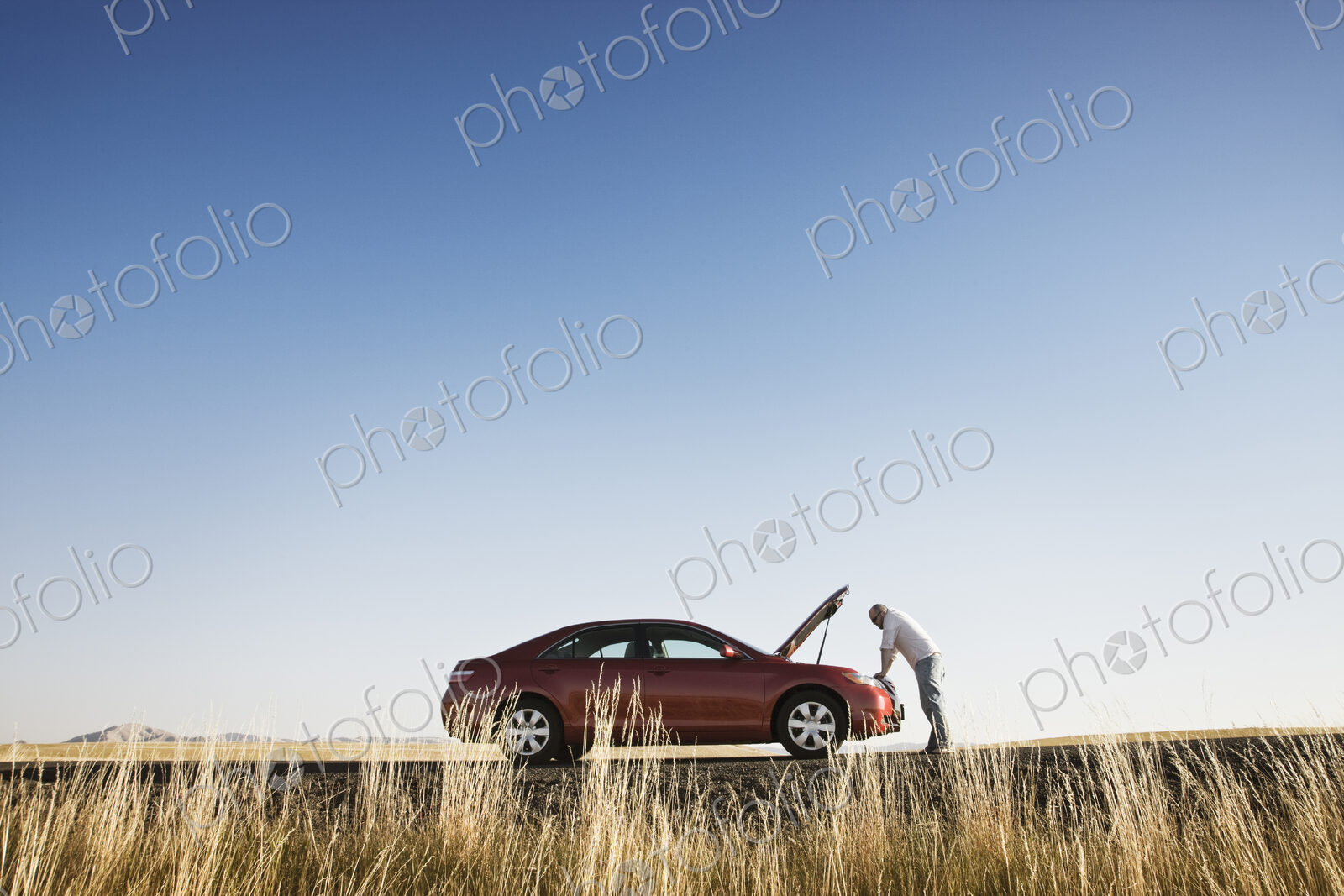A man looking in the engine compartment of his parked car parked along a highway