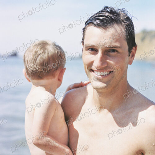 A man on the beach, smiling, carrying his young son A man on the beach, smiling, carrying his young son