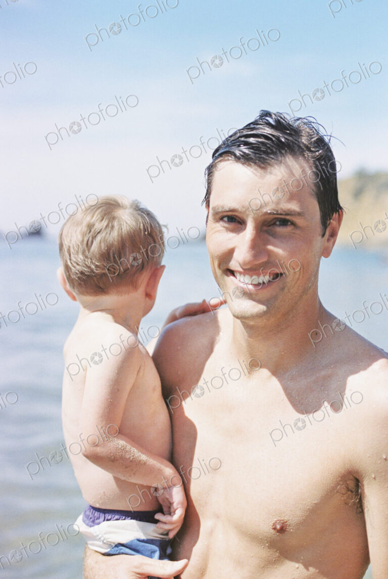 A man on the beach, smiling, carrying his young son