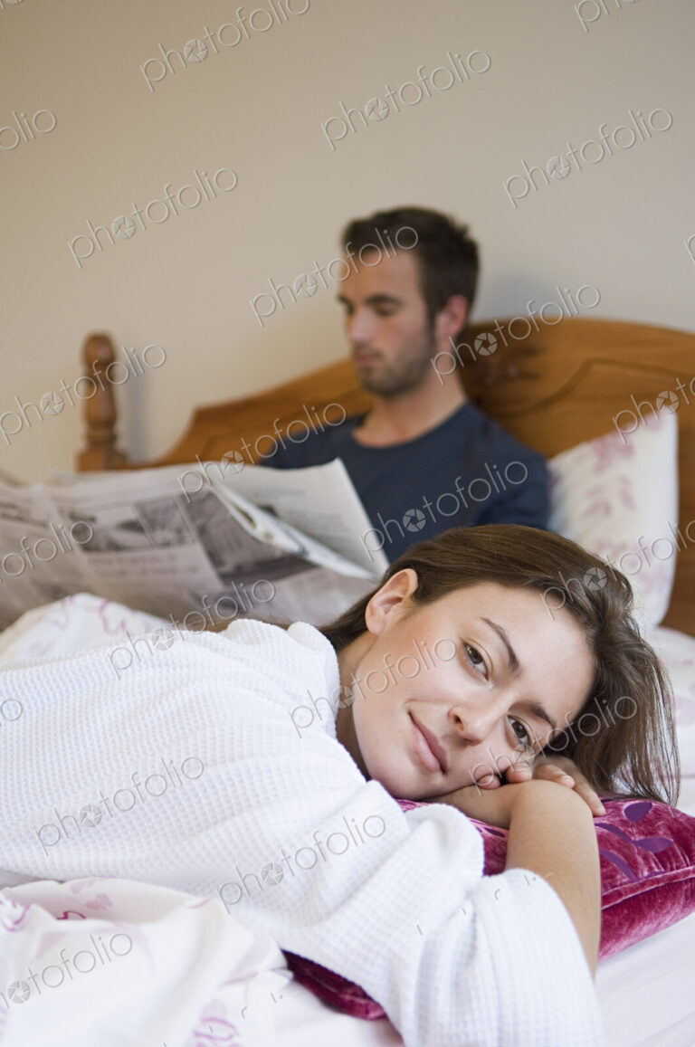 A man reading the paper in bed, and a woman relaxing smiling.