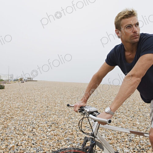 A man riding a bicycle on a shingle beach.